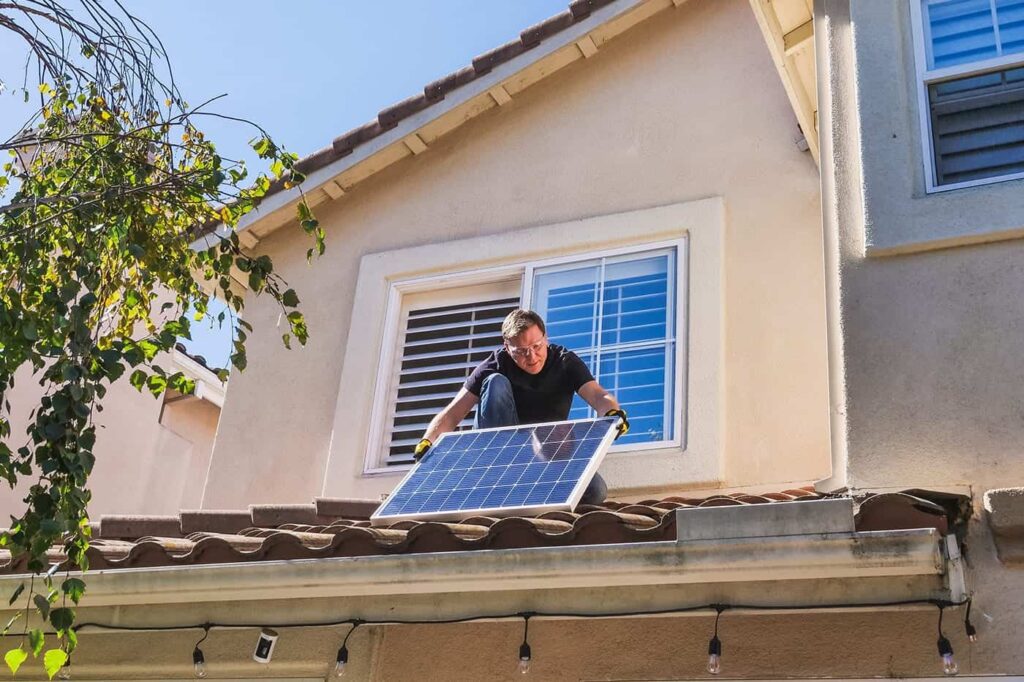 Man installing solar panel on a terracotta roof, reaching through the window